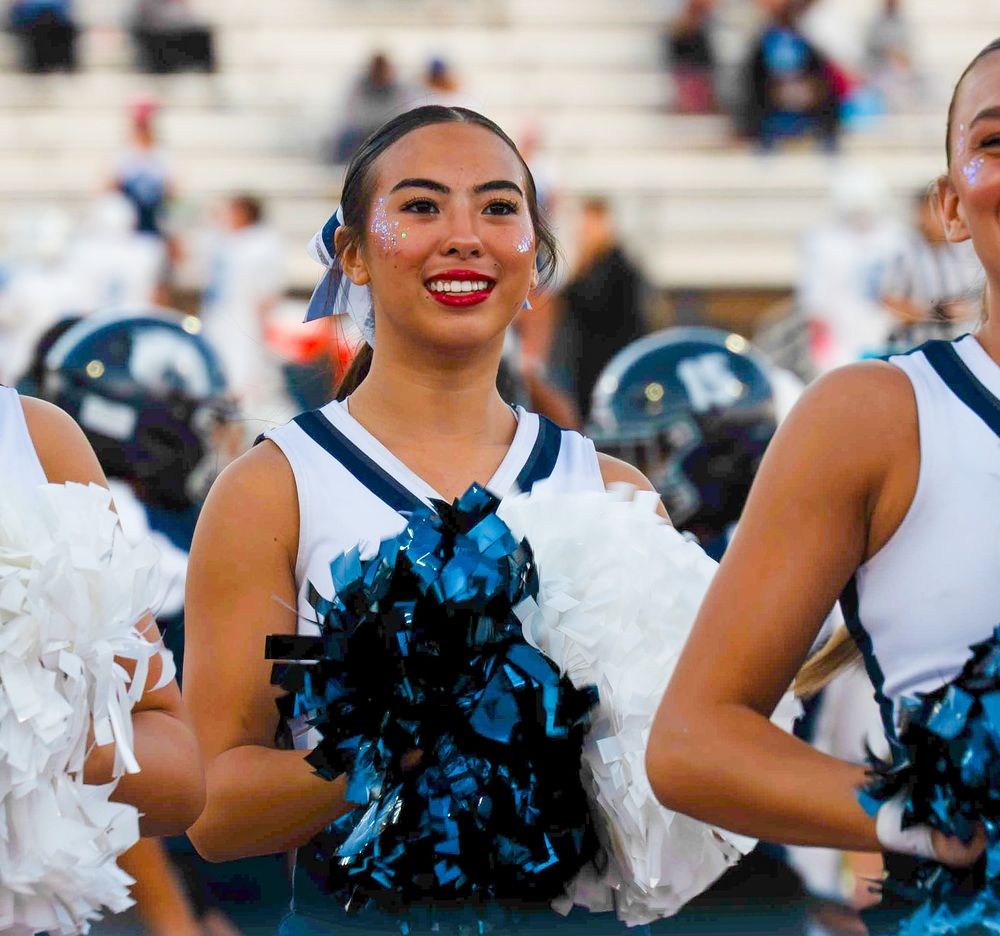 Cheerleader smiling with blue and black pom-poms at a high school football game.