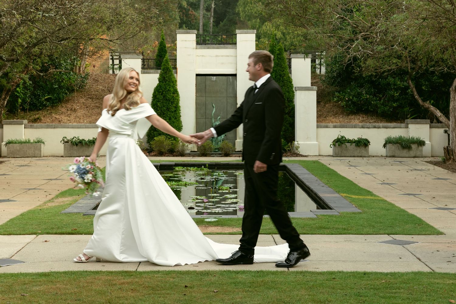 Wedding couple walking hand in hand by reflecting pool in formal garden setting.