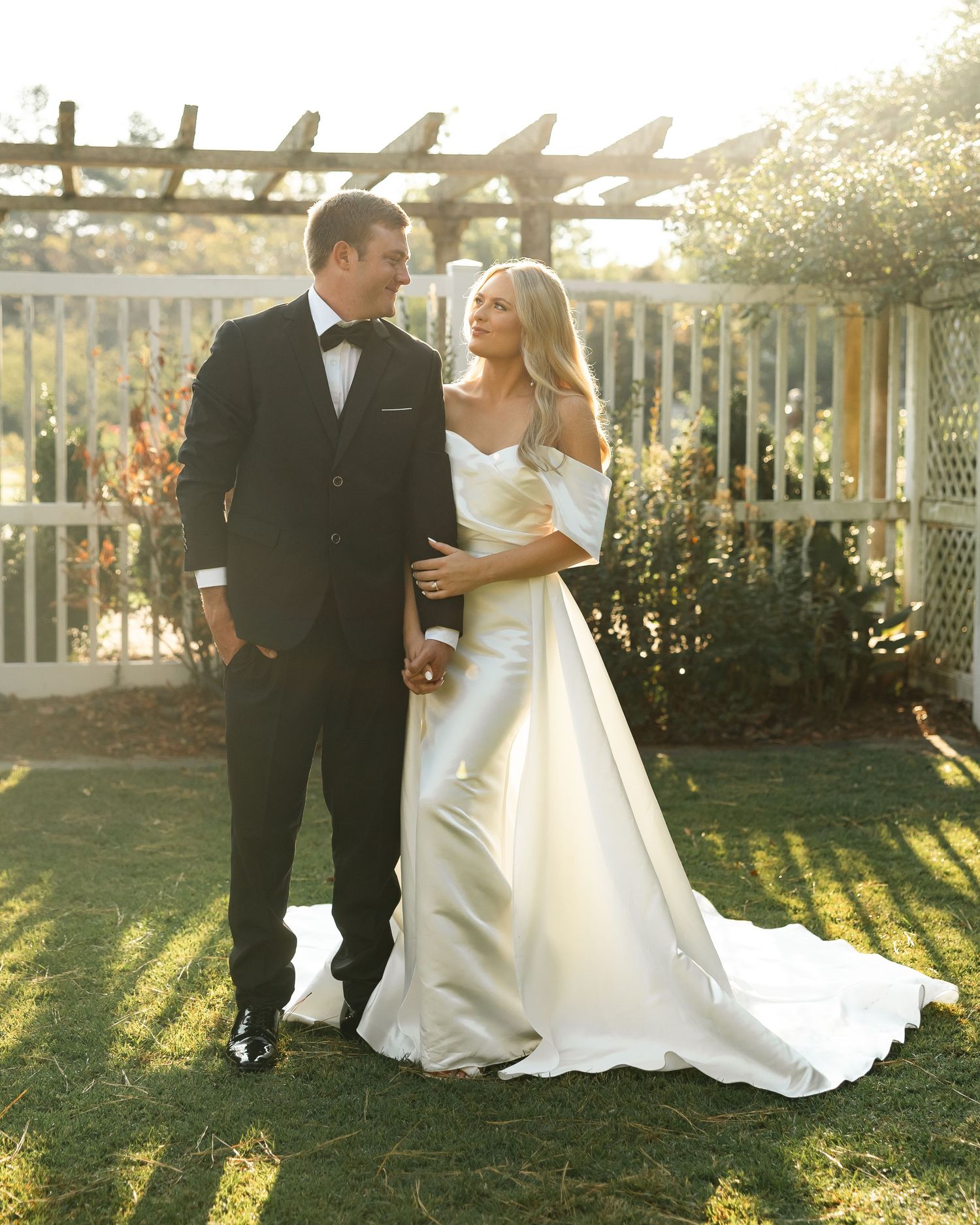 Couple in formal wedding attire posing in garden setting during golden hour.