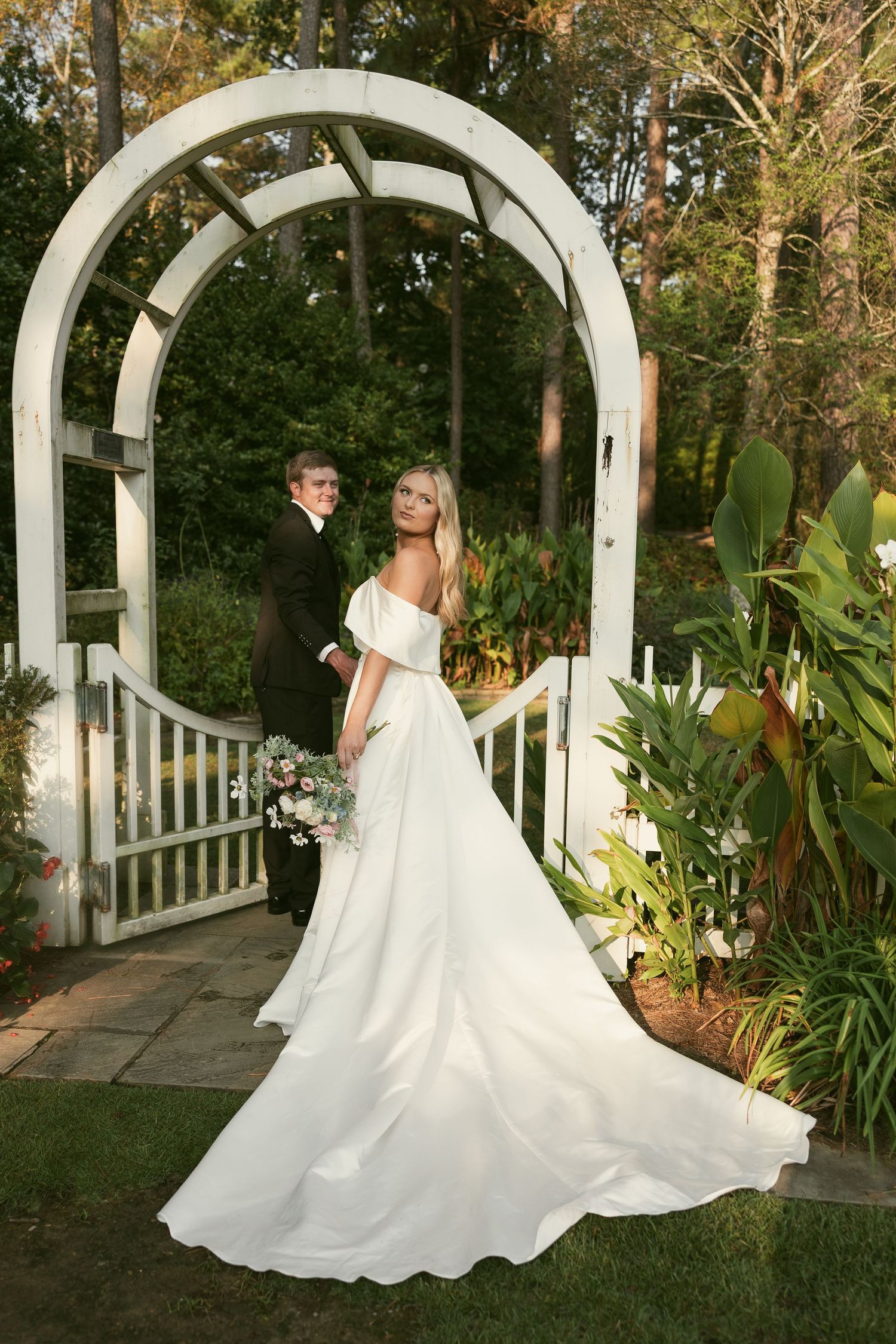 Elegant wedding dress with flowing train photographed under white garden arch surrounded by lush greenery.