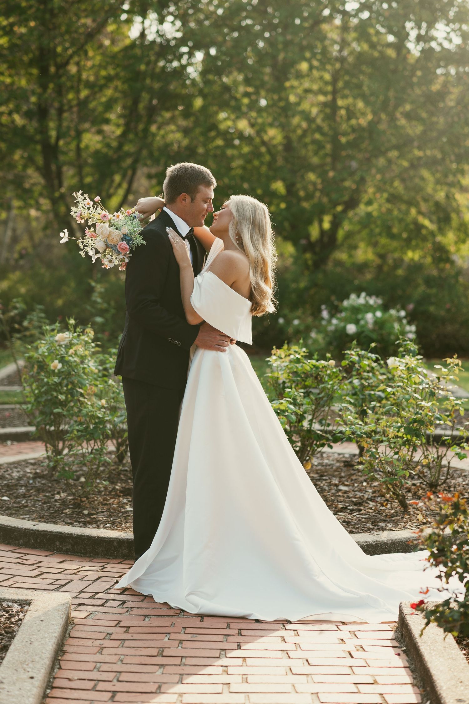 Romantic wedding couple embracing on brick pathway surrounded by garden flowers at sunset.