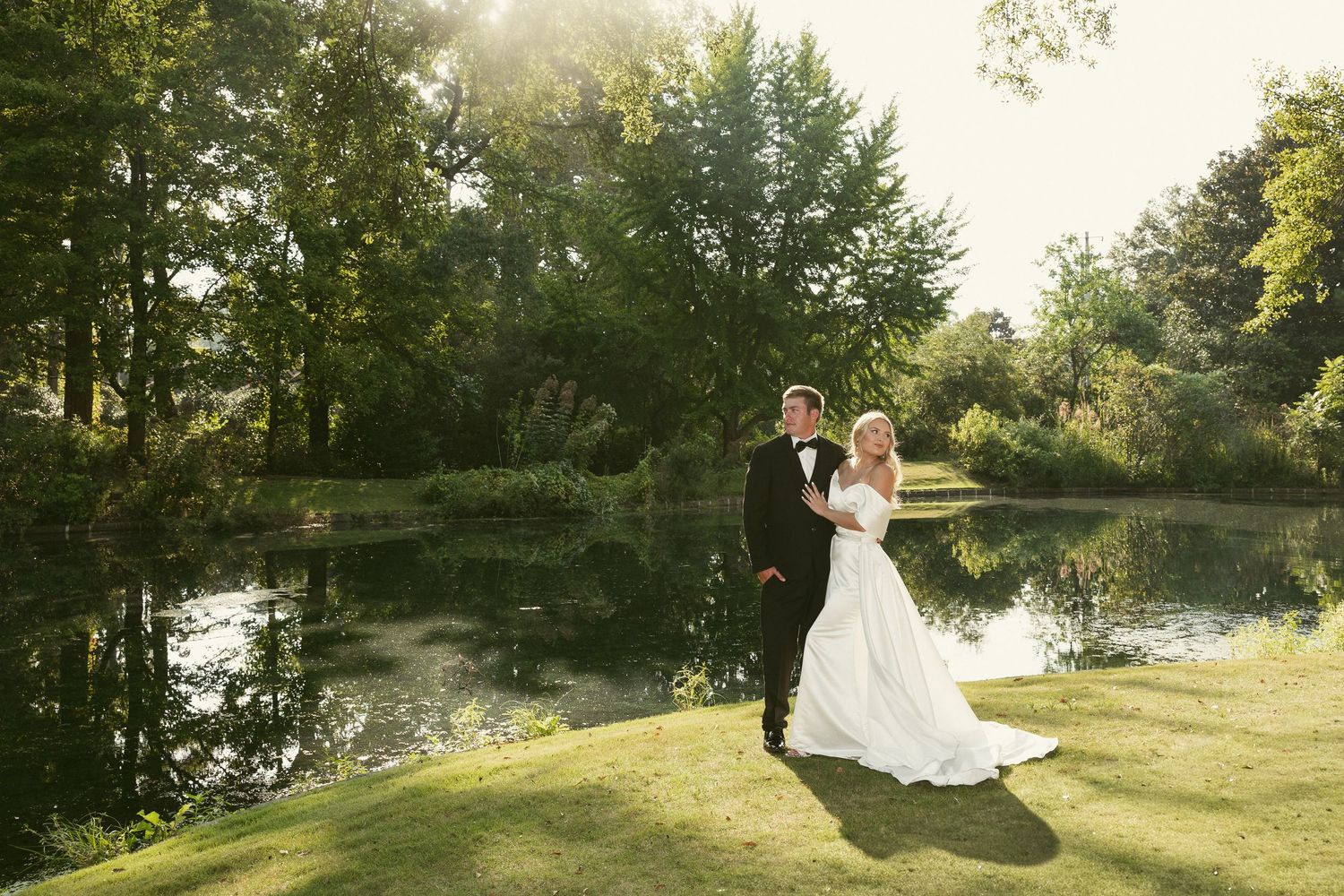 Wedding couple poses by tranquil pond during golden hour in landscaped garden.