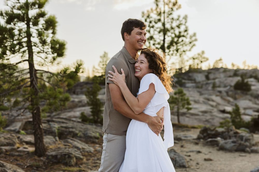 Romantic couple embracing outdoors among pine trees and rocky terrain.