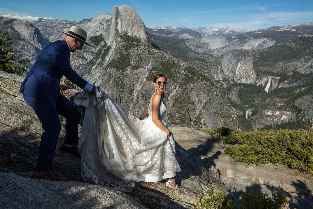 Two people in wedding attire stand at Glacier Point overlook with Half Dome and Yosemite Valley in the background.