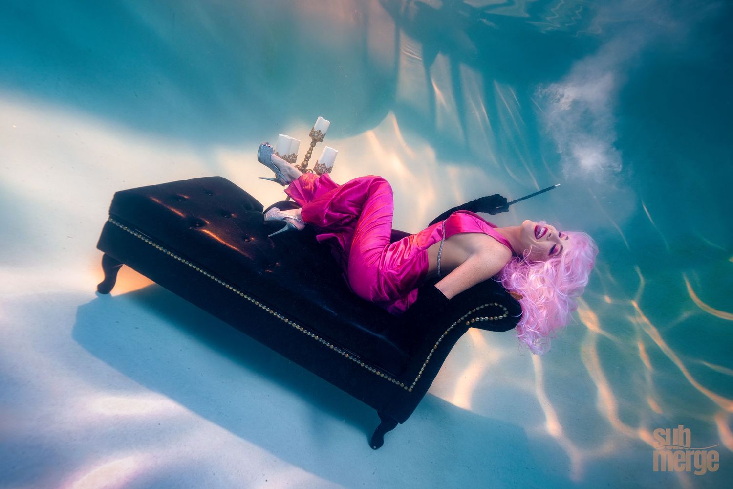 A figure in a pink dress lies on a black piano in an underwater photography scene, surrounded by shimmering blue reflections.