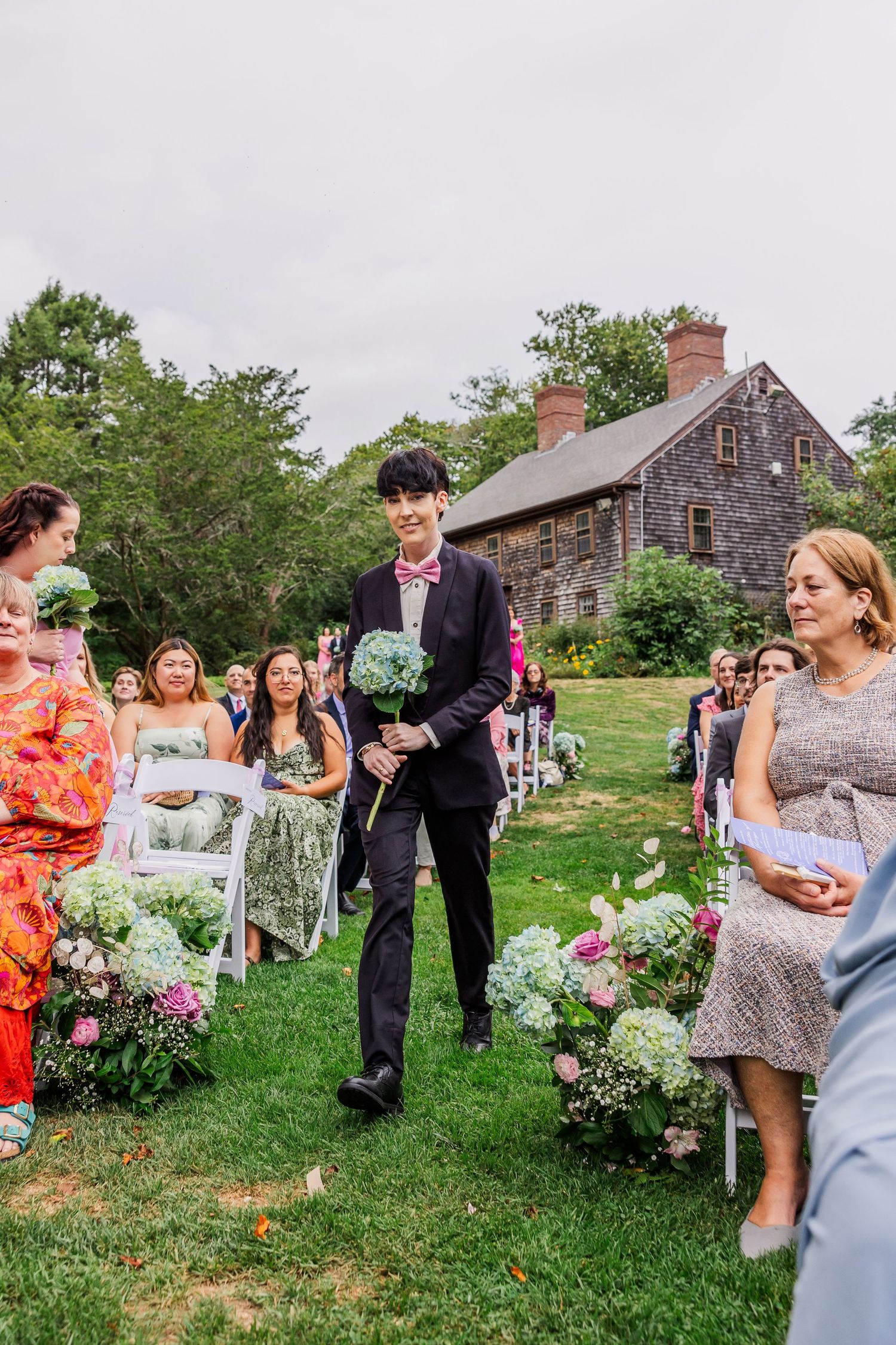 Outdoor wedding ceremony at rustic stone house with floral aisle decorations and guests gathered on the lawn.
