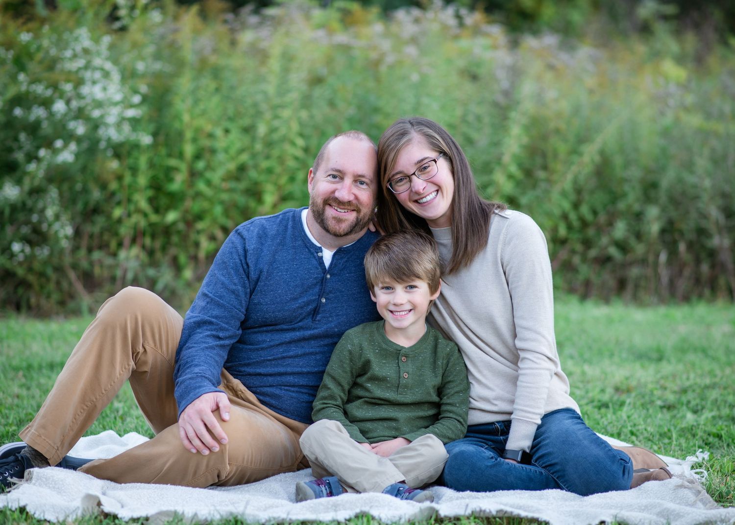 Three people sitting together on a blanket outdoors with green foliage in the background.