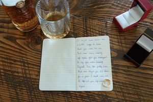 Handwritten wedding vows on paper next to a whiskey glass and ring box on wooden table.