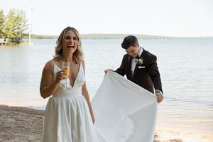 A couple playfully holds a white wedding dress veil on a beach with water and trees in the background.
