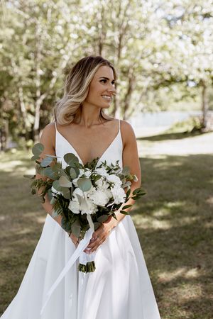 Bride in elegant white dress holds beautiful bouquet of white flowers and eucalyptus outdoors.