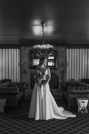 Black and white wedding photograph showing a bride in an elegant white gown standing in front of a stone fireplace.