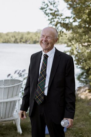 A gentleman in a dark suit and striped tie stands near white Adirondack chairs by the water.