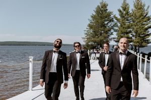 Groomsmen in dark suits walk along a waterfront dock on a sunny day.