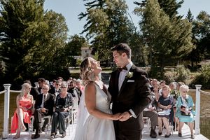 Wedding ceremony guests gathered outdoors under trees watching couple exchange vows on sunny day.