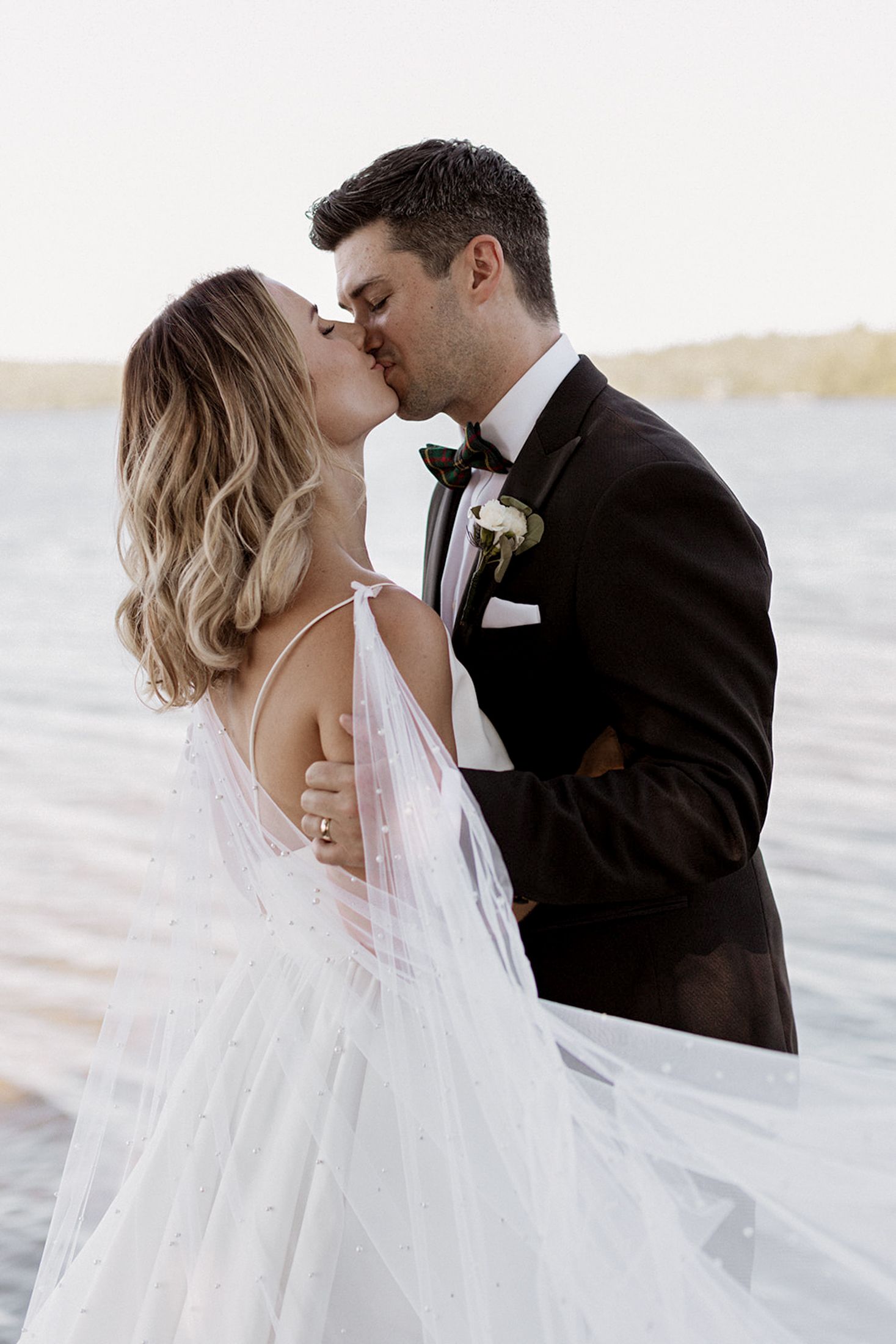 A romantic beach wedding couple embraces as the bride's veil flows in the ocean breeze at sunset.