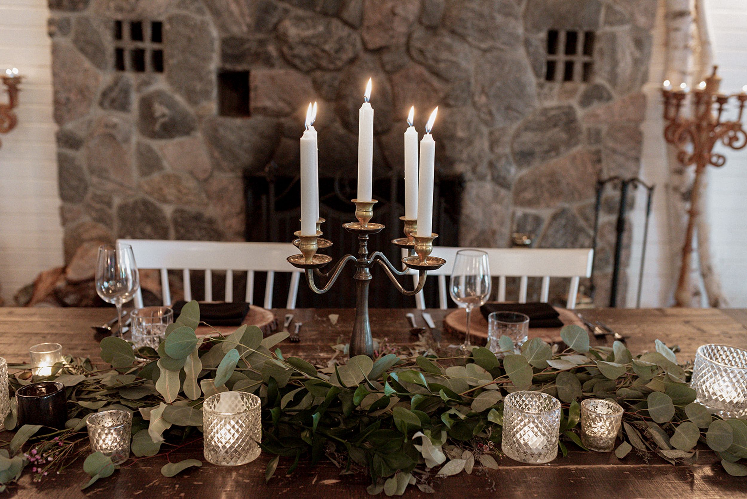 White candles in glass holders line a rustic stone wall with greenery and twinkling lights creating an elegant ambiance.
