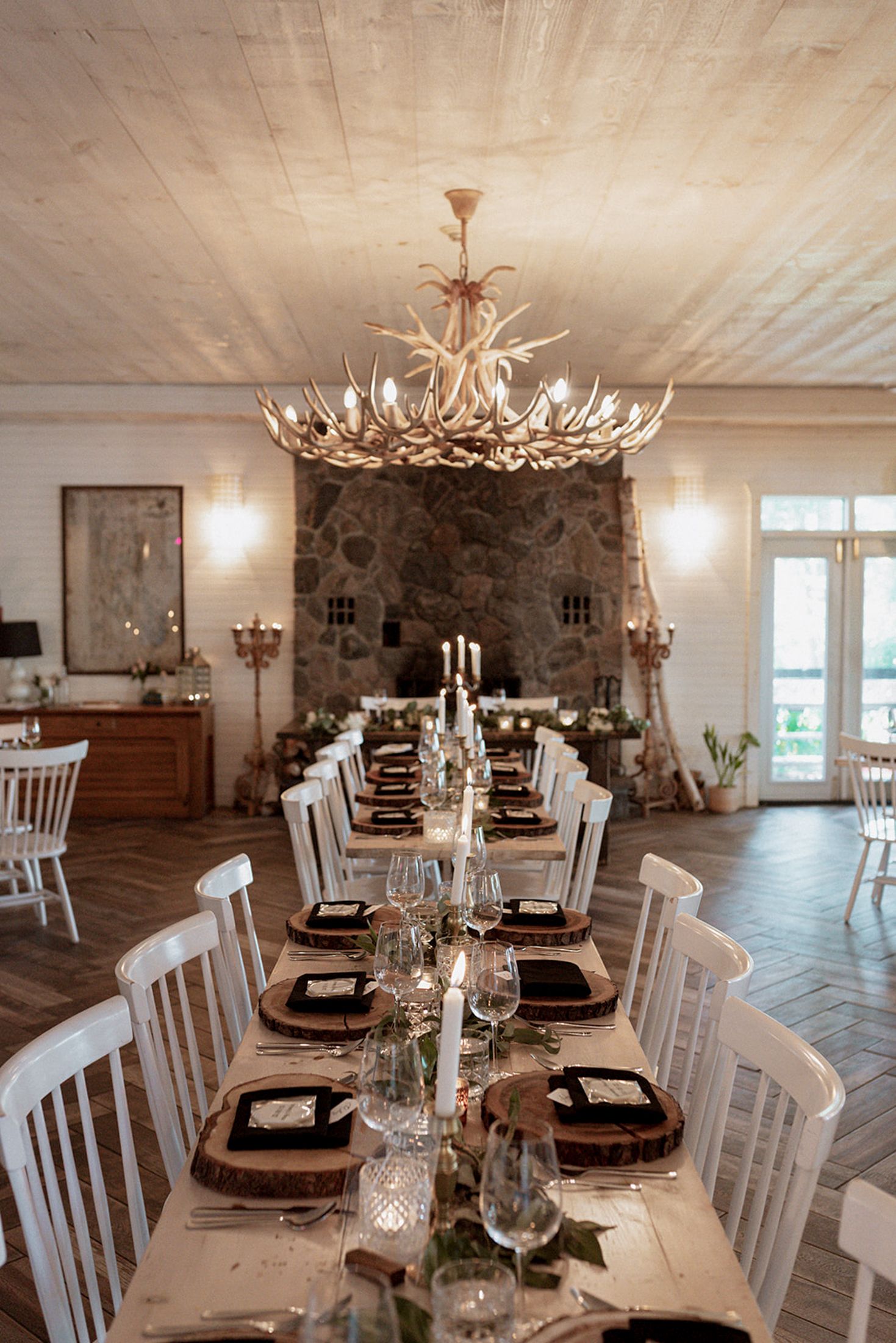 Rustic dining room with white chairs, antler chandelier, and stone walls showcases elegant table settings and natural lighting.