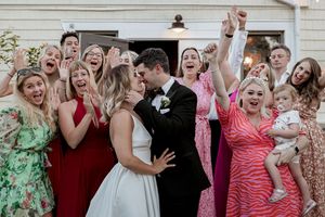 Wedding party celebrates joyfully outside a white building in colorful dresses.