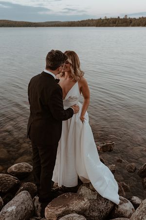 Couple in wedding attire shares romantic moments by a lake with rocky shoreline.