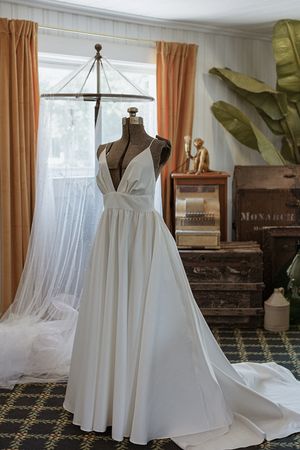 Elegant white wedding dress displayed on mannequins in a sunlit room with peach curtains.