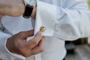 Close-up of hands adjusting a gold monogrammed cufflink on a white dress shirt sleeve.
