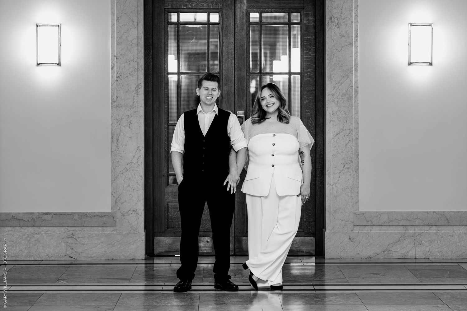 A black and white photograph of a couple in formal attire standing in an elegant hallway with wall sconces.