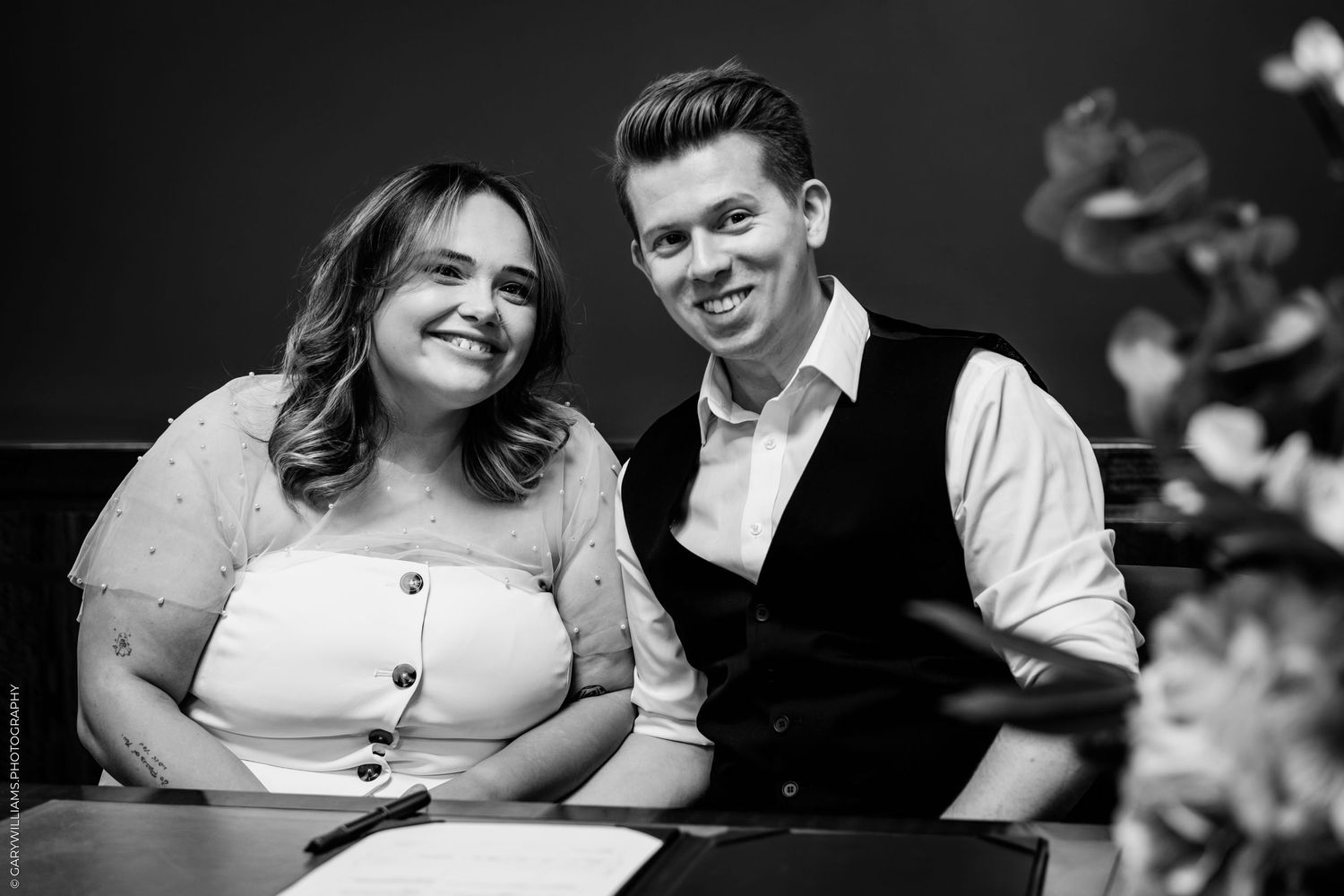 A black and white portrait of a couple sitting together and smiling, with the person on the right wearing a vest.