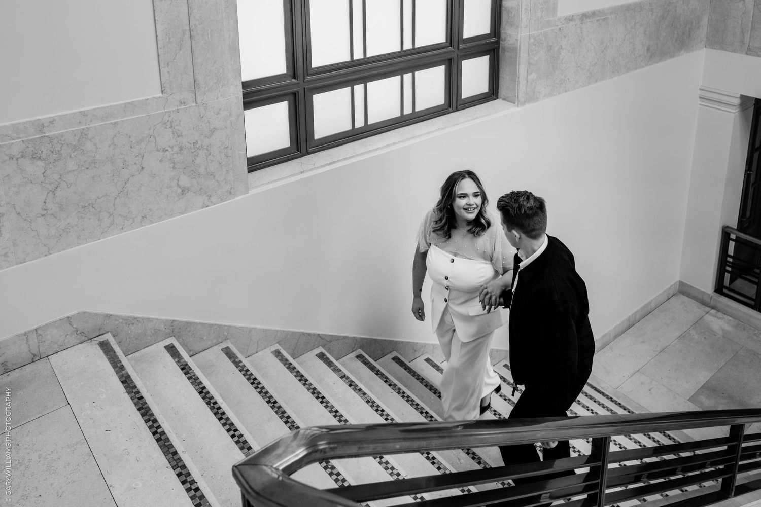 A couple in formal attire stands together on a staircase landing in a black and white photograph.