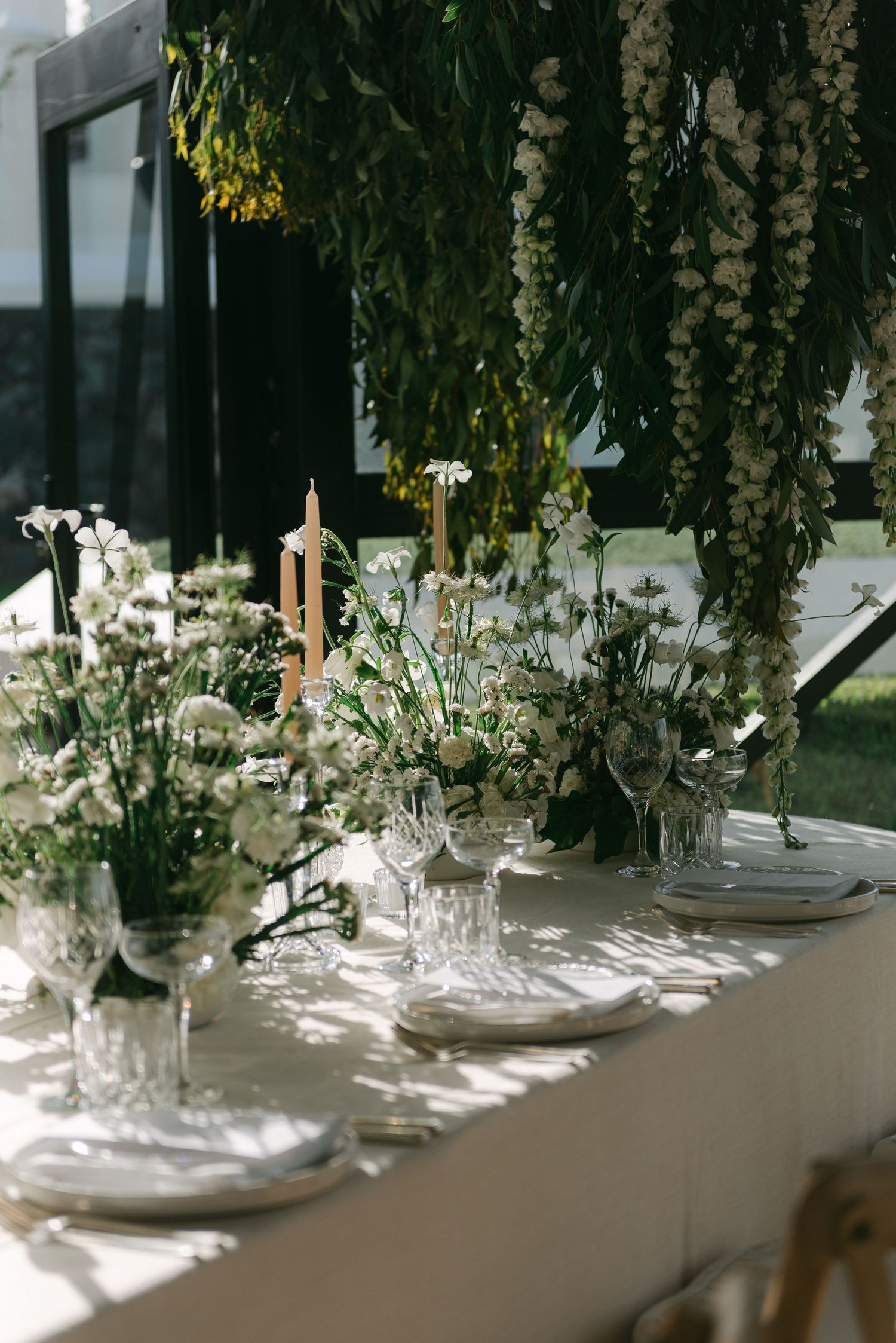 Formal table setting with crystal glassware and white floral centerpieces under hanging floral arrangements.