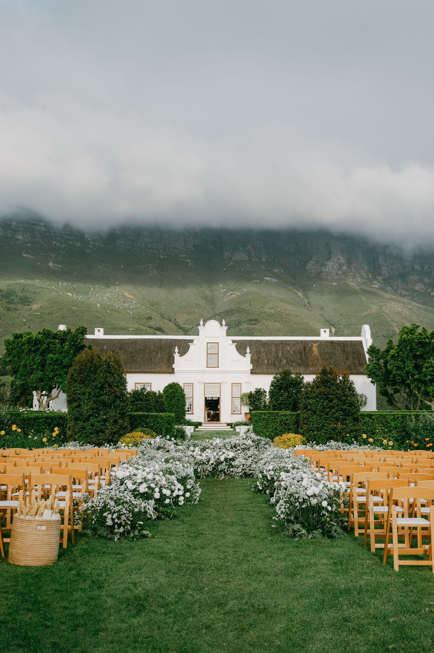 Elegant outdoor wedding ceremony setup with white chairs and floral arrangements leading to a colonial-style white building.