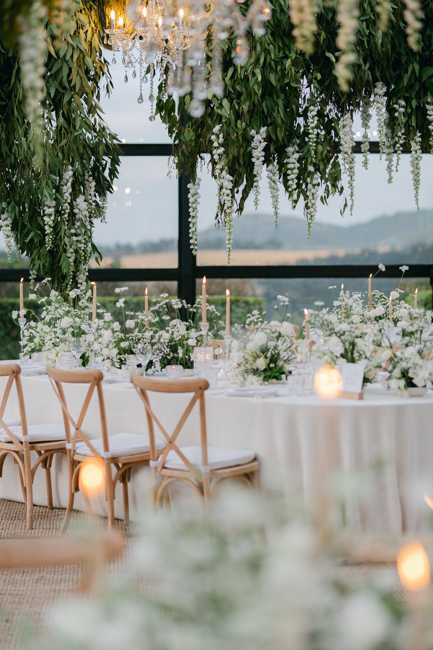 Elegant wedding reception setup with hanging floral arrangements and wooden chairs under soft candlelight.