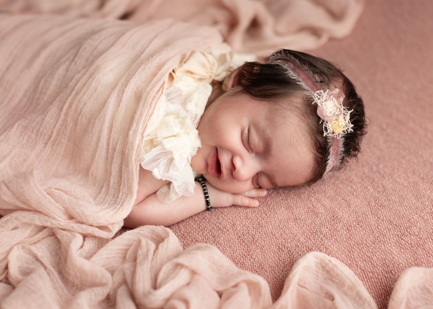 Sleeping newborn baby in soft pink blanket with delicate lace details and floral headband accessory.