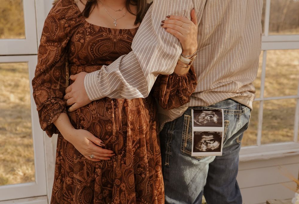 A couple holds ultrasound photos while wearing a brown paisley dress and striped shirt.
