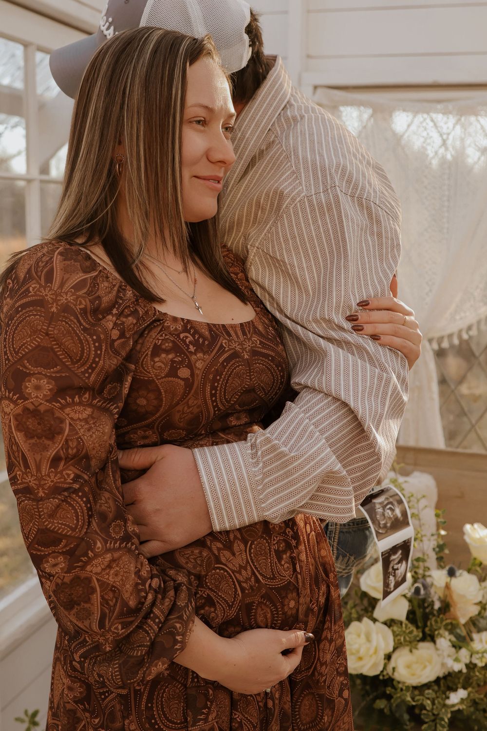 Intimate maternity moment showing brown paisley dress and striped shirt against sunlit window.