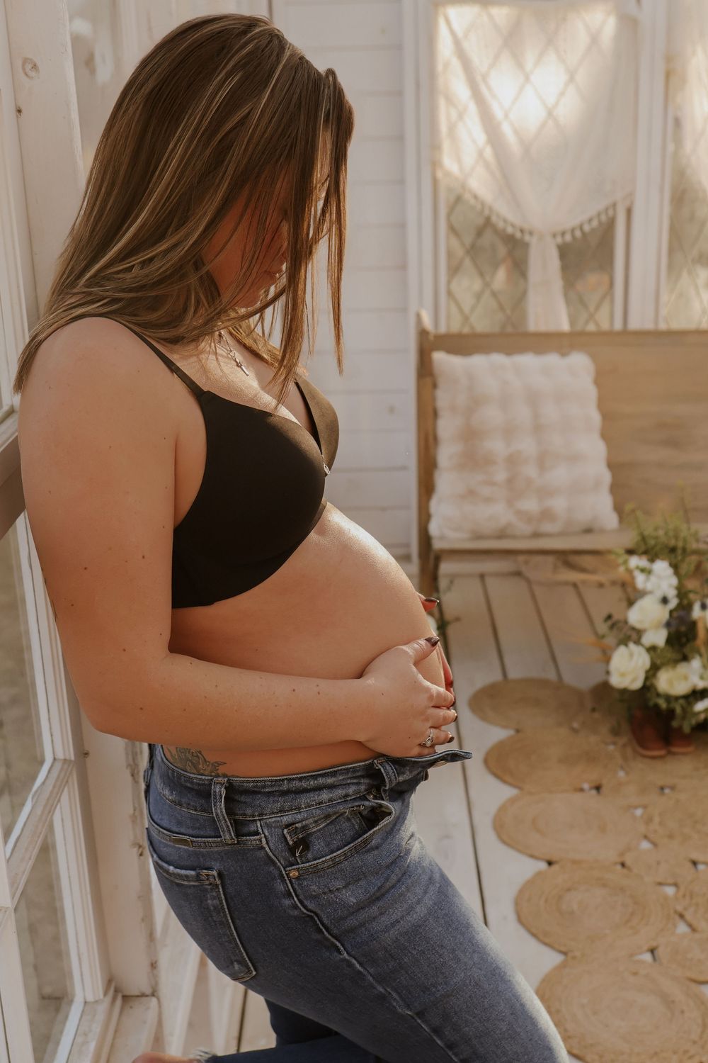 A pregnancy announcement photoshoot showing a person in jeans and black top holding their belly in natural light.