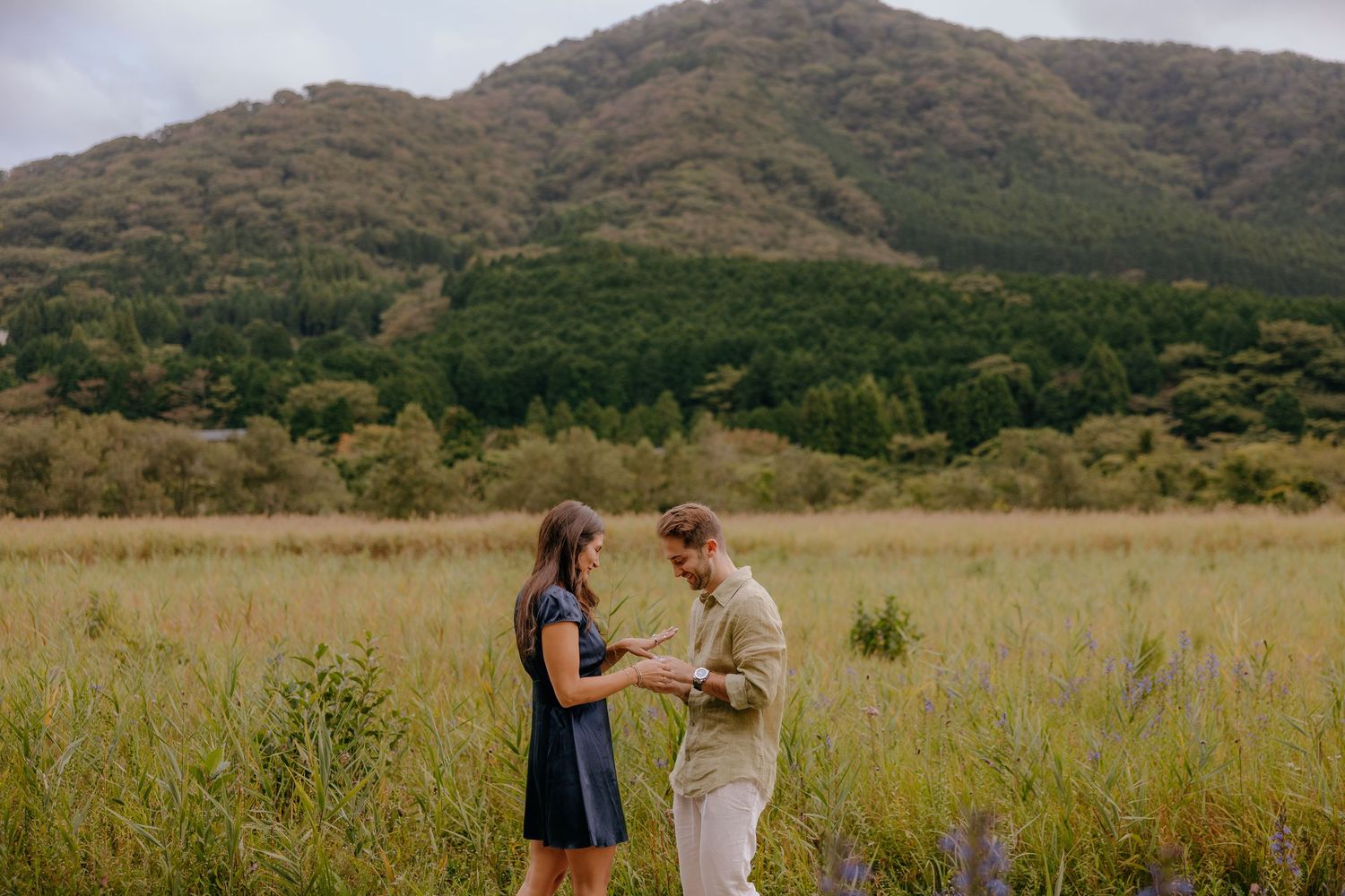 Two people stand together in a golden grass field with mountains and trees in the background during sunset.