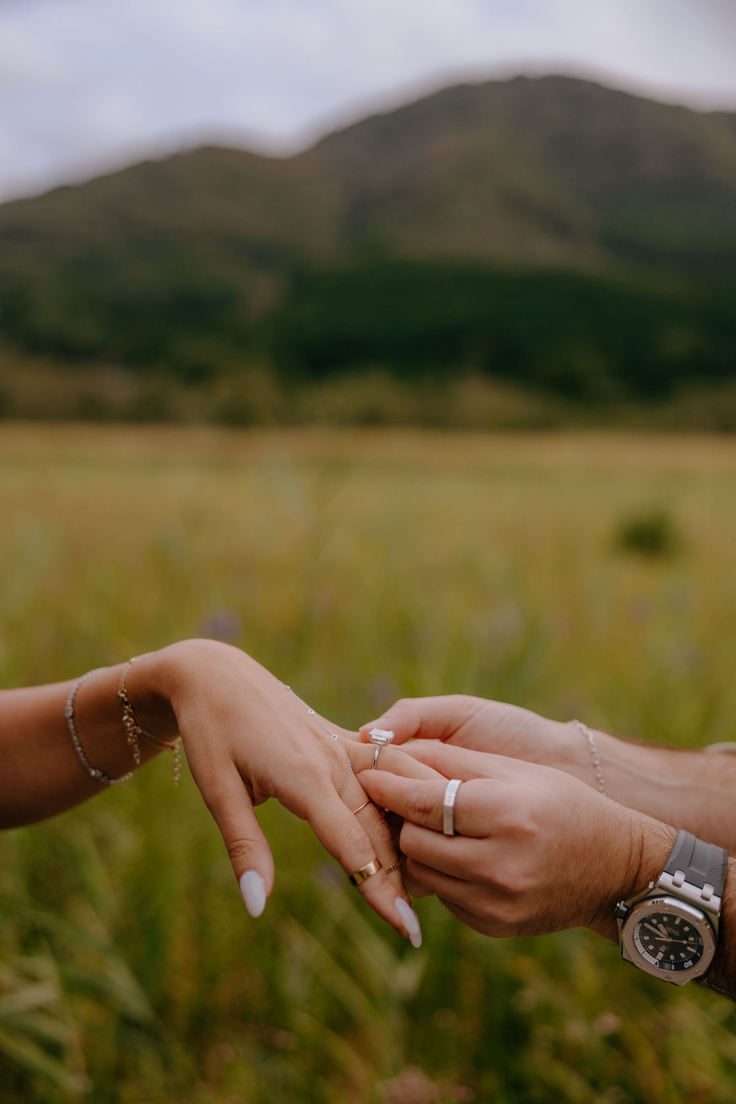 Two hands reaching towards each other wearing rings and watches against a blurred mountain landscape background in Hakone Japan