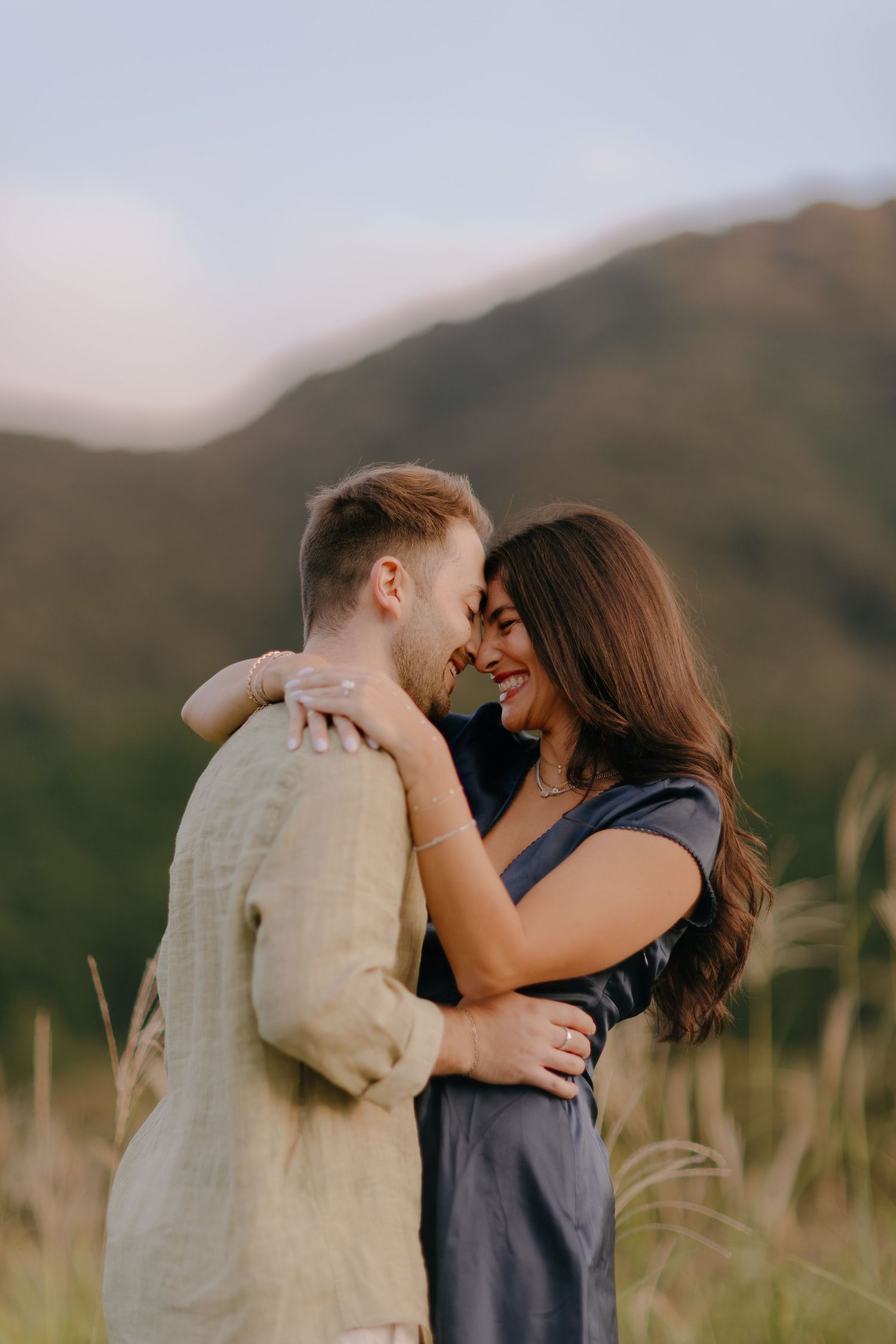A romantic couple embraces and kisses in a field with mountains in the background at sunset.