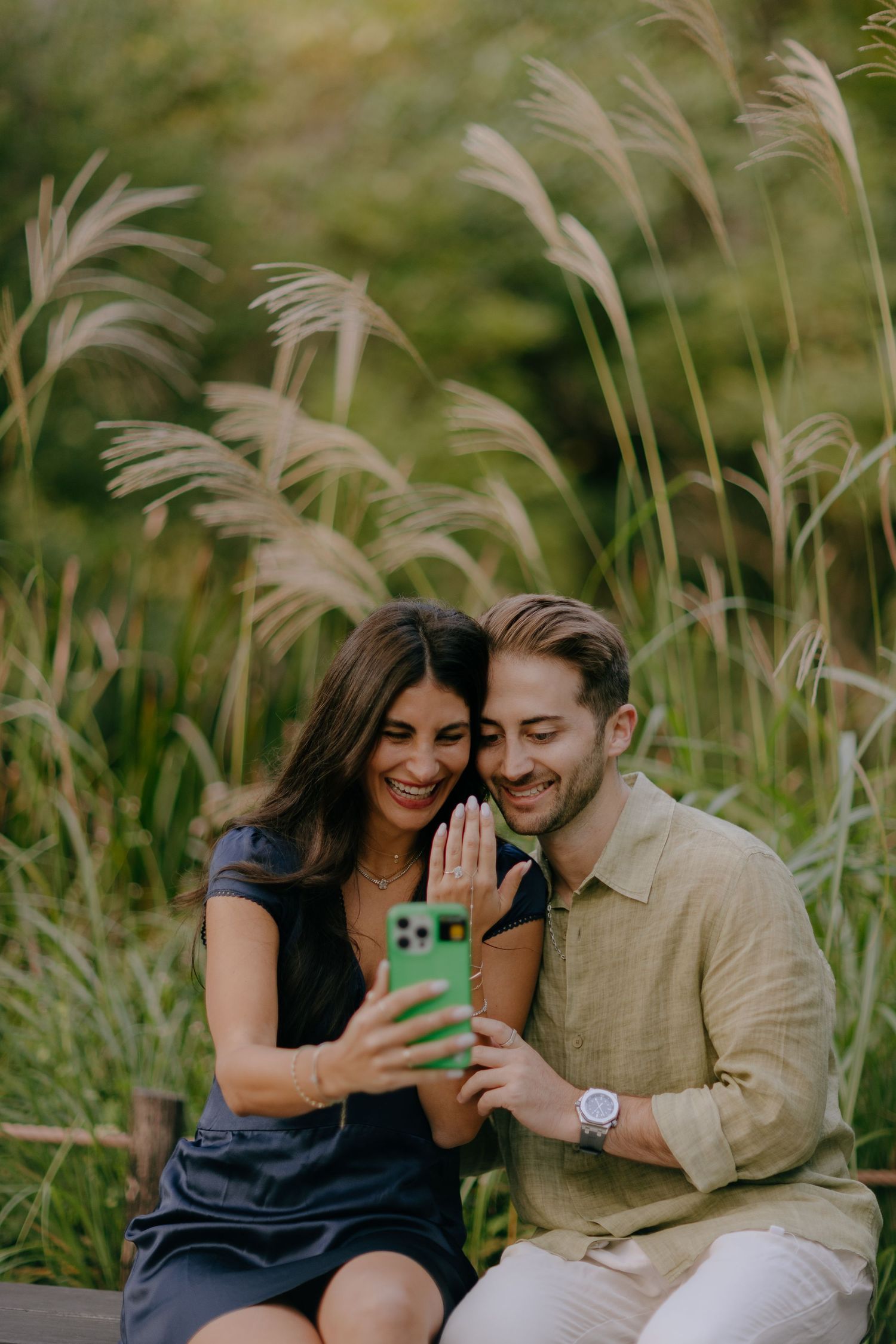 A couple sits together in tall grass and pampas plumes while sharing joyful moments and displaying an engagement ring at Hakone Japan