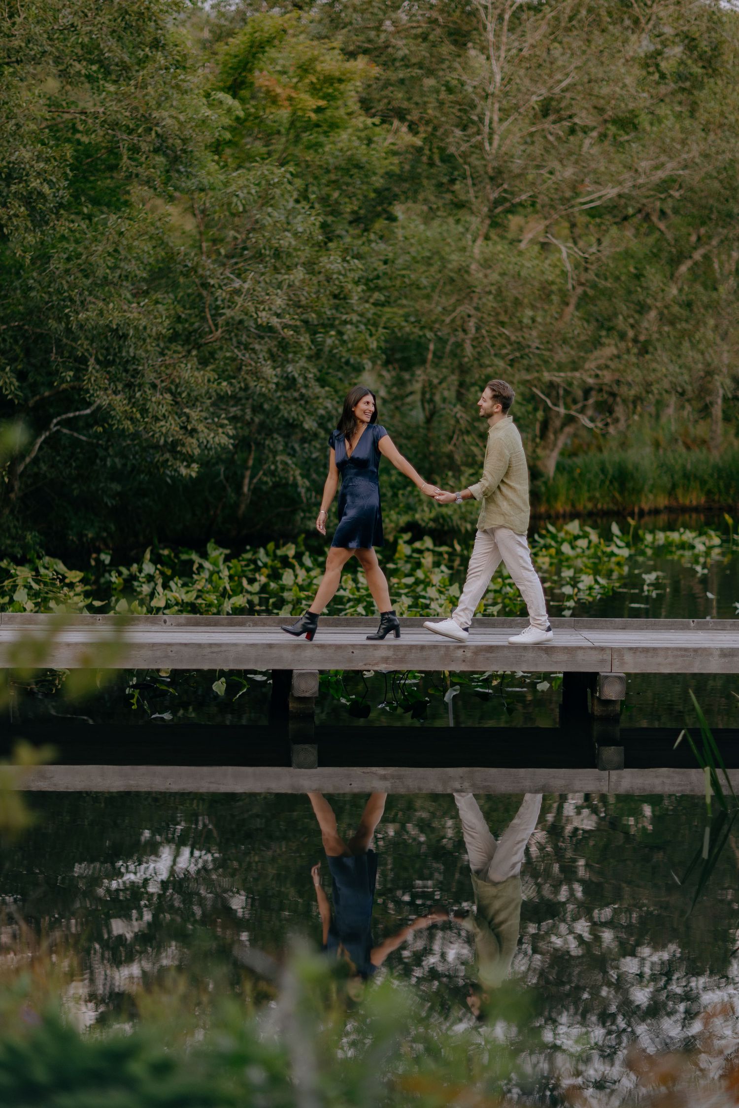 Two people sit and walk along a wooden boardwalk over calm reflective water surrounded by lush greenery at Hakone Japan
