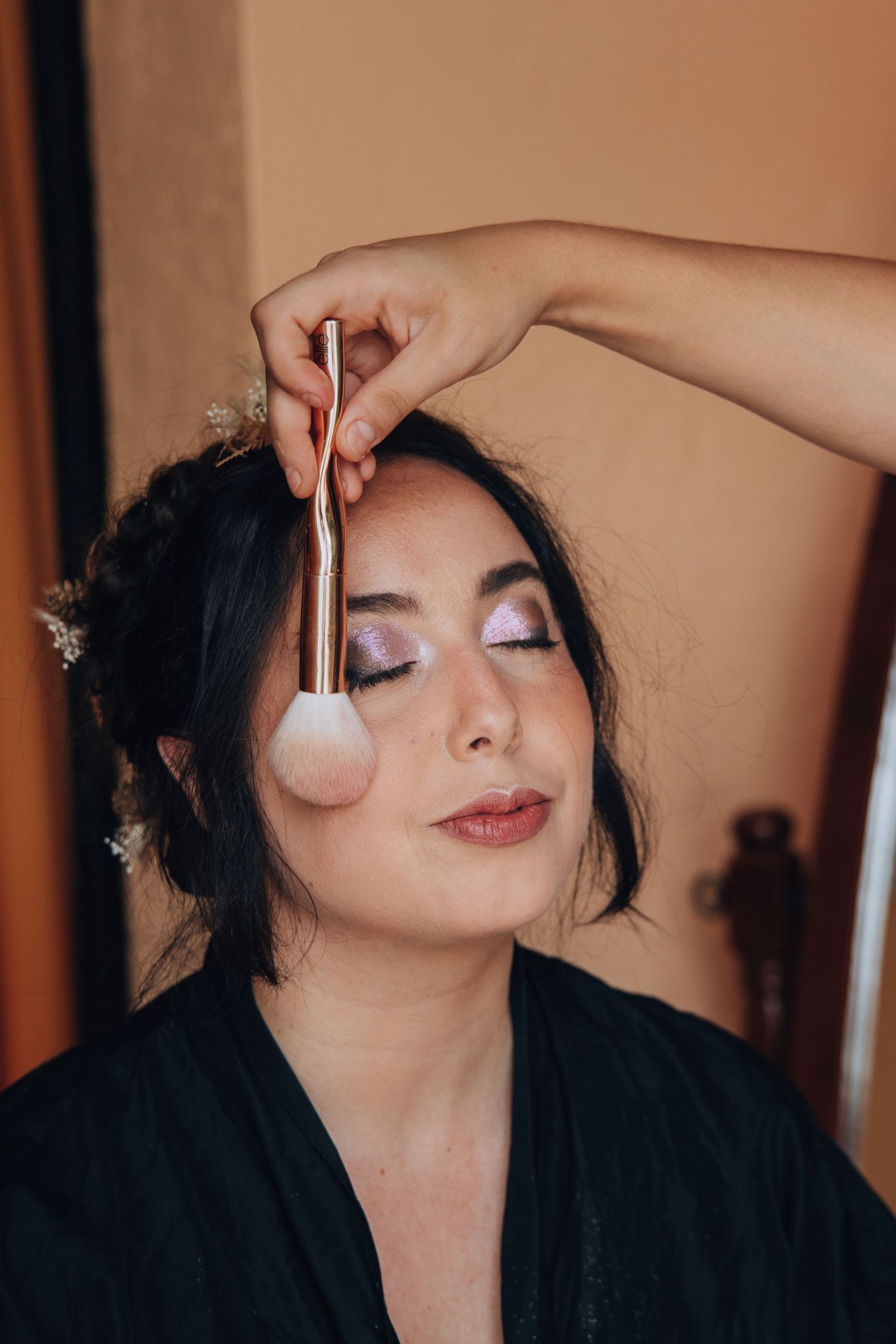 Bride with floral hair accessories gets makeup applied with large brush before wedding.