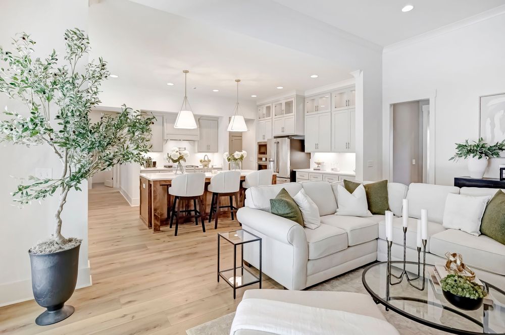 Open concept living space with white furniture, hardwood floors, and natural light streaming through large windows.