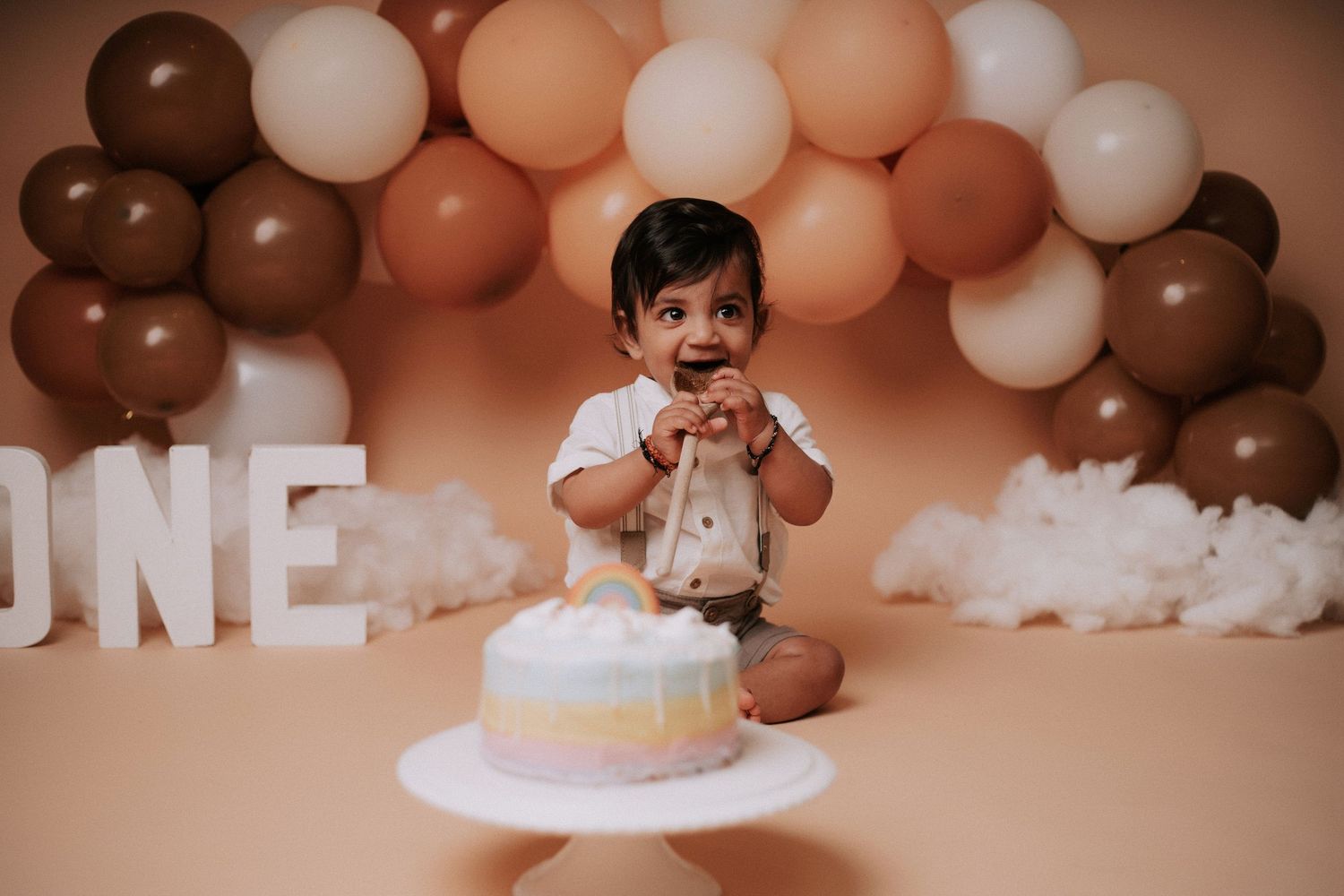 Cake smash photo session with brown and peach balloon backdrop and white cake stand setup.
