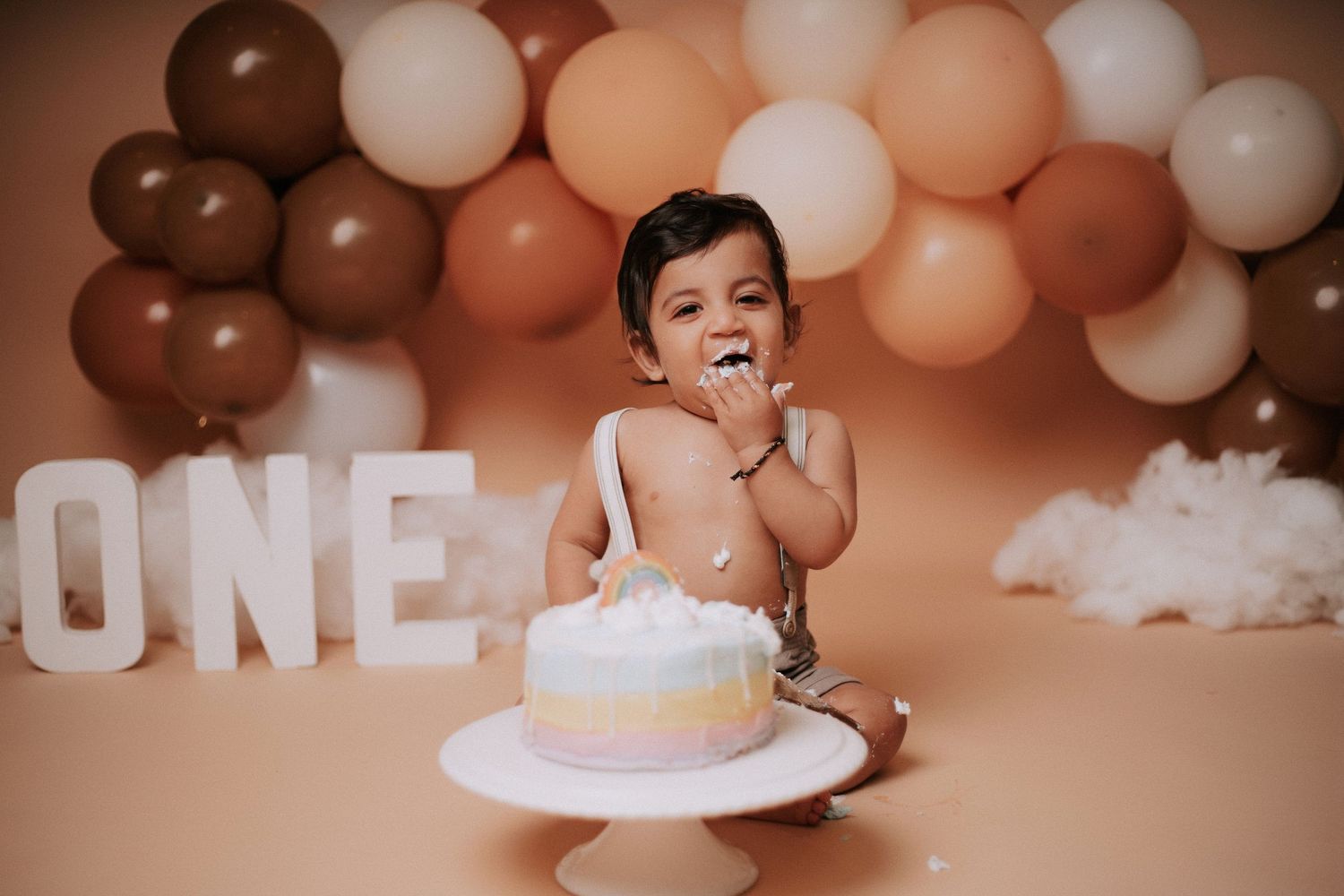 First birthday setup with neutral toned balloon arch and white cake stand against warm backdrop.