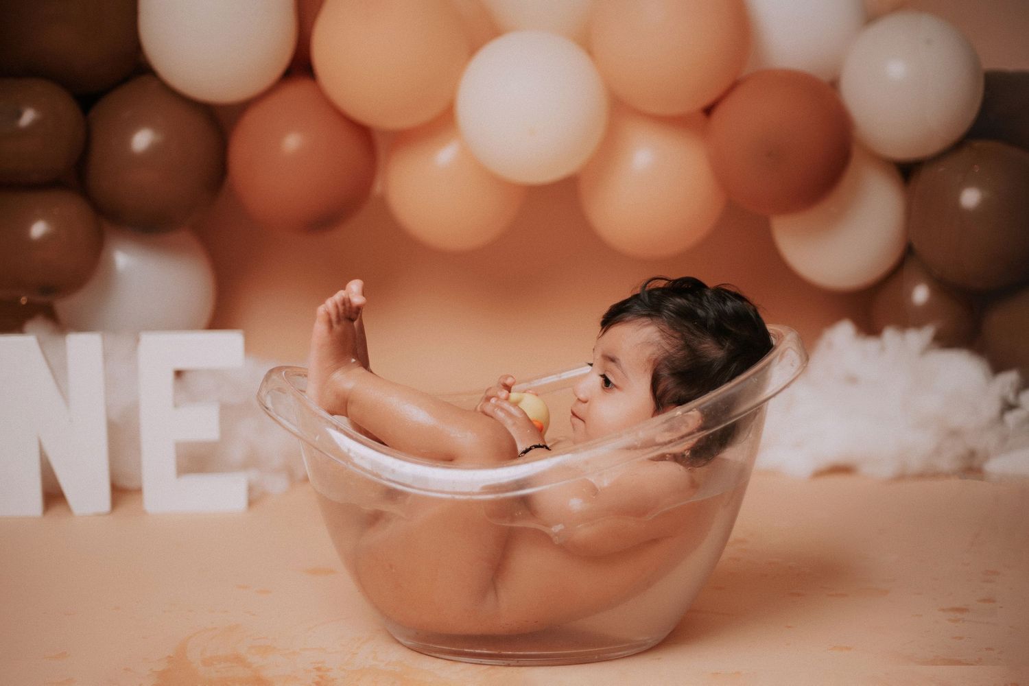 Baby photography session featuring transparent tub and decorative balloon arrangement in warm tones.