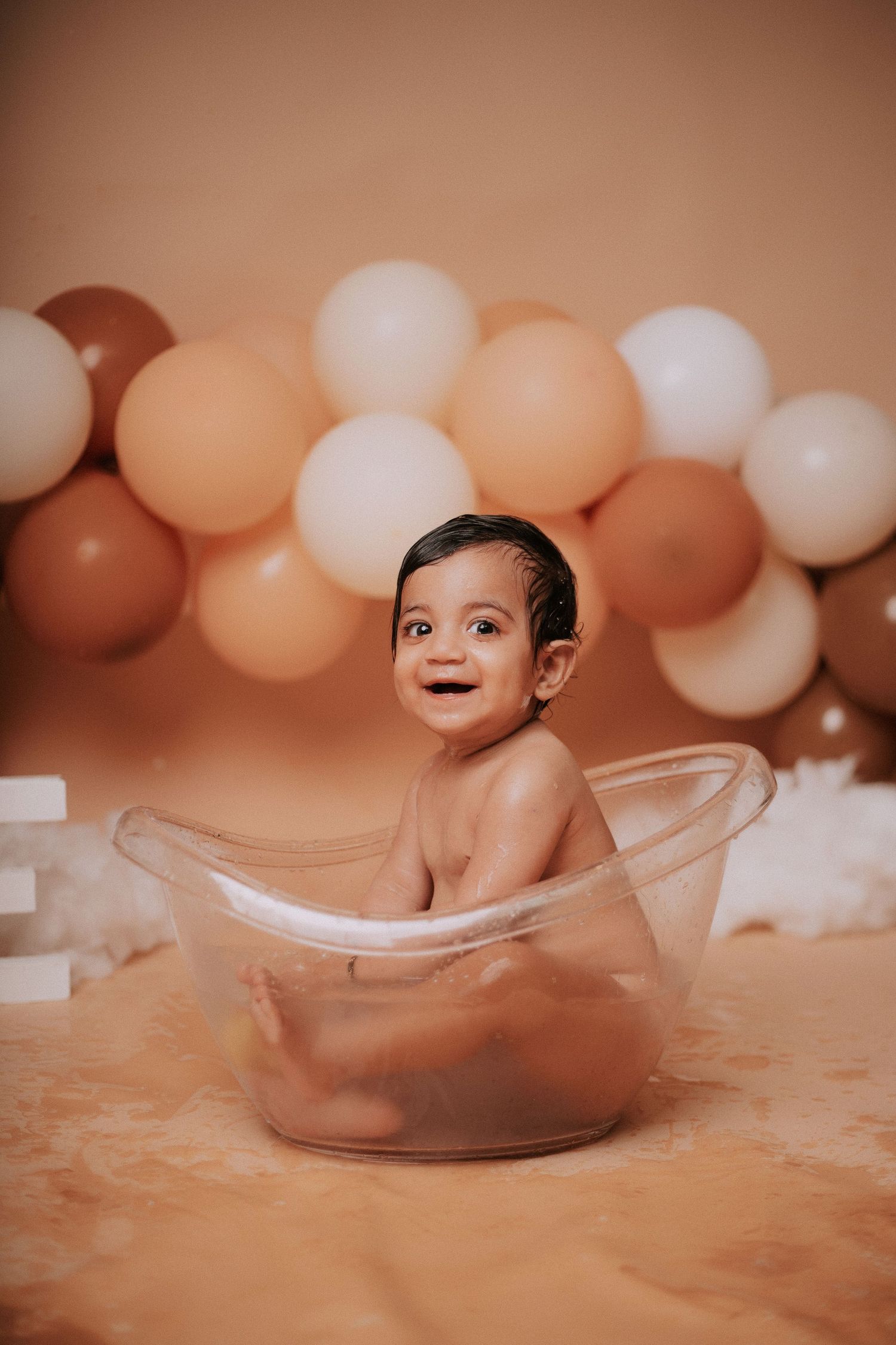 Artistic cake smash photo with clear bathtub prop and balloon garland in neutral colors.