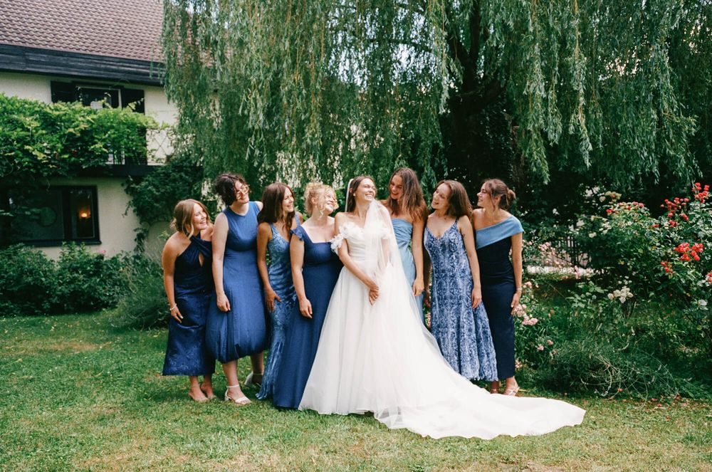 A bridal party in coordinating blue dresses poses together under a weeping willow tree.