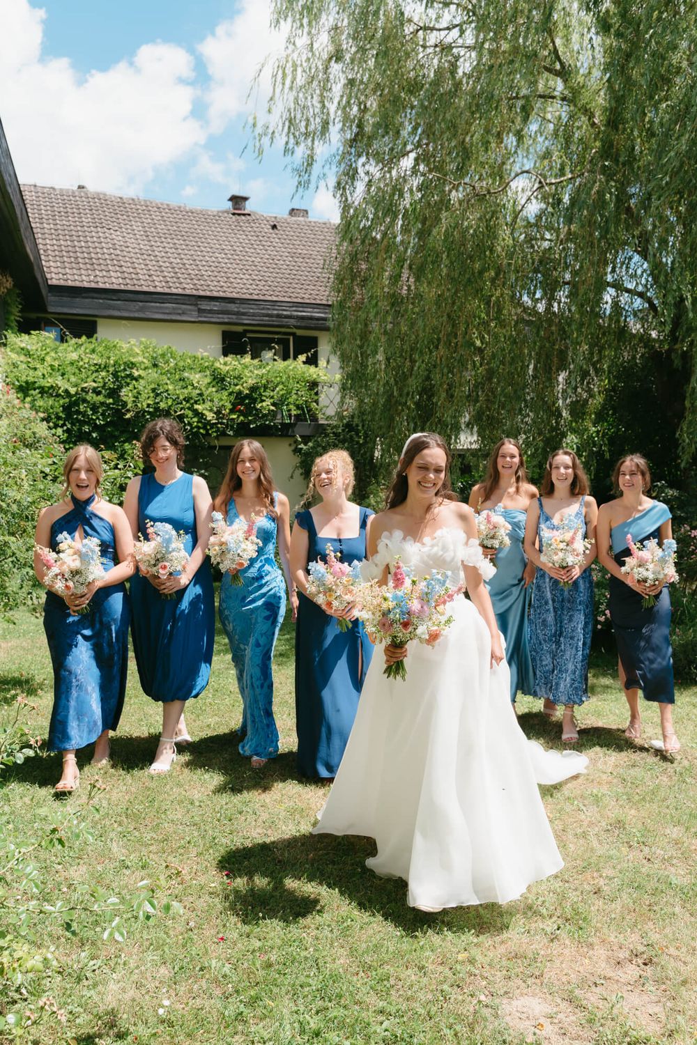 Bridal party in blue dresses and white gown pose together outdoors under willow trees on sunny summer day.