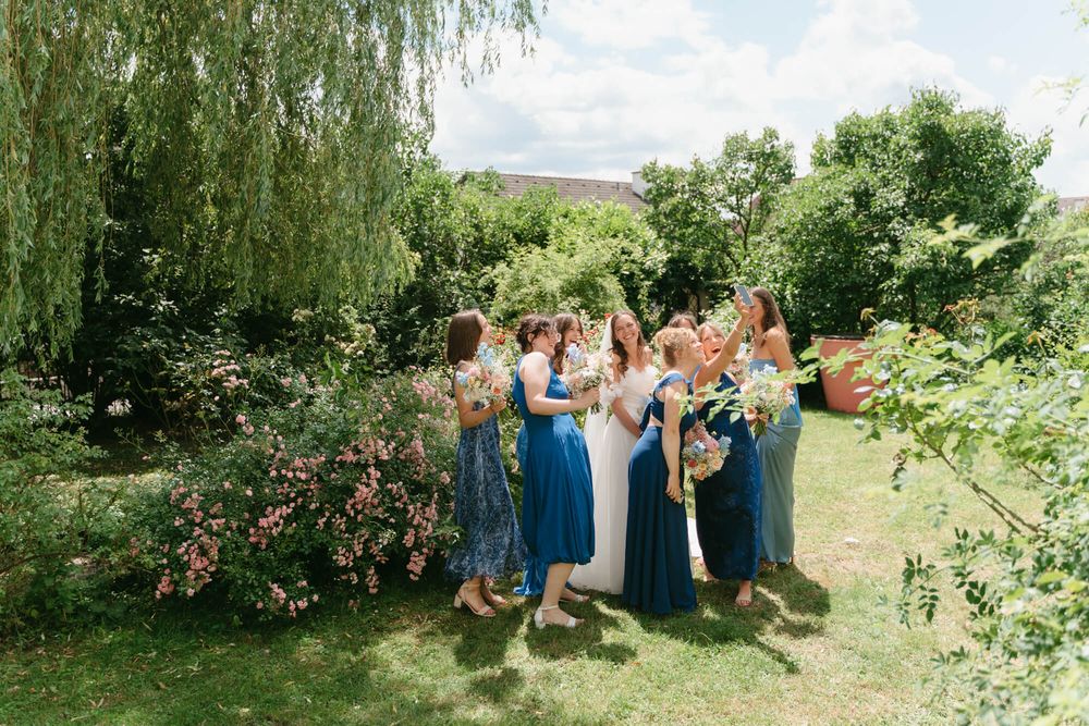 Wedding party stands in a lush garden setting with weeping willow trees and natural greenery on a sunny summer day.