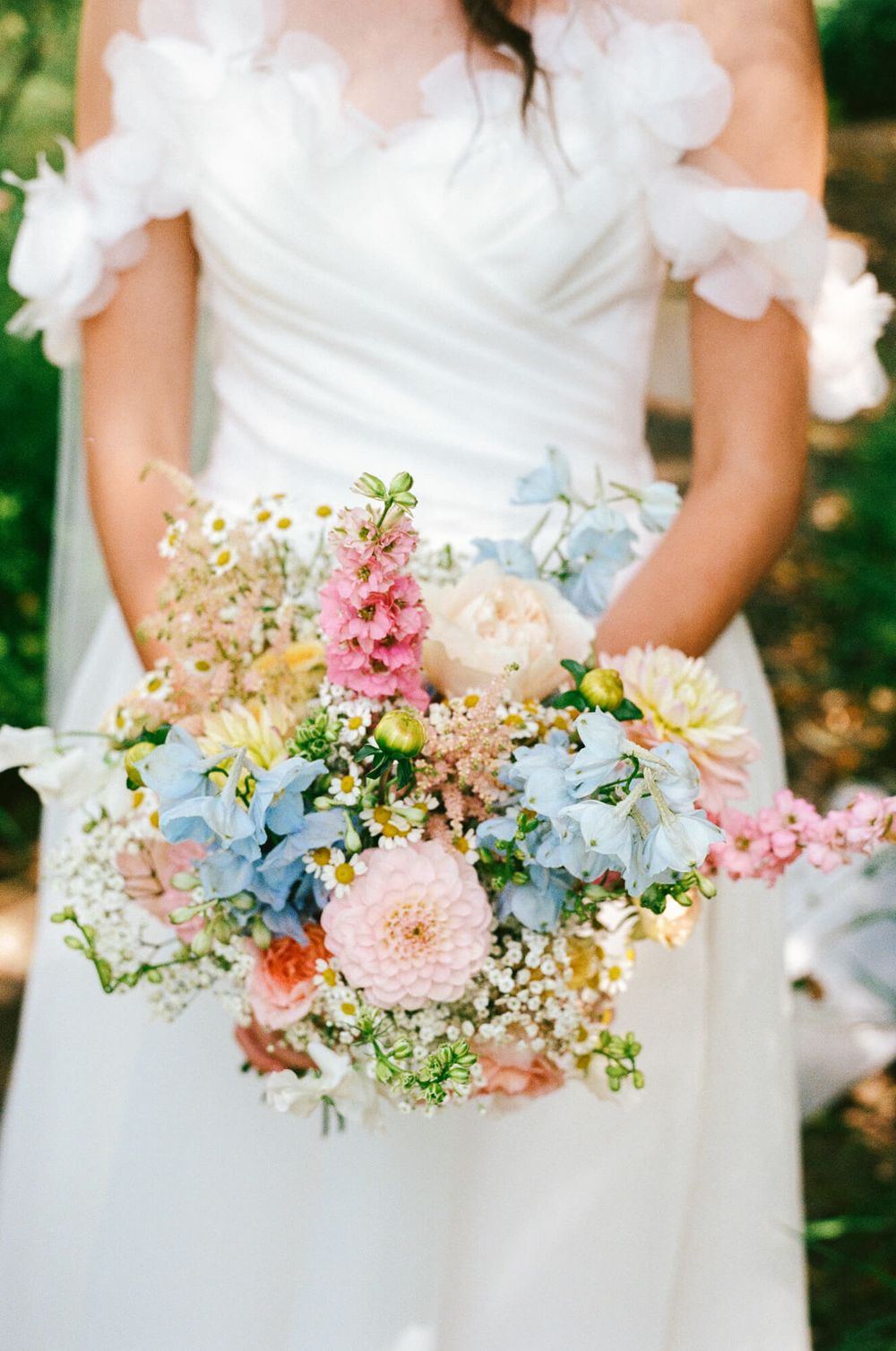 Bridesmaids hold pastel bouquets with pink dahlias, blue hydrangeas and wildflowers against white dresses at outdoor wedding.